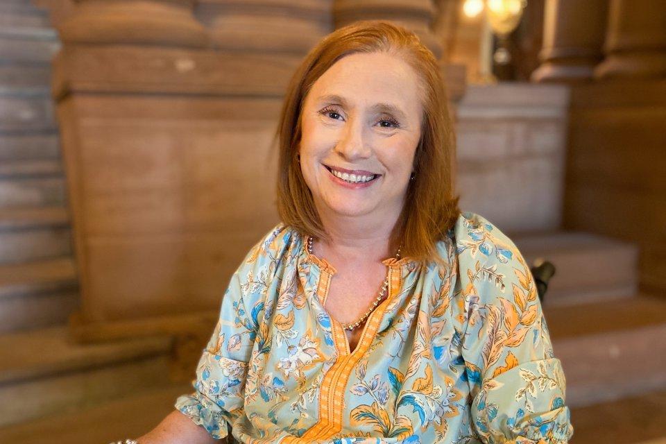 Kimberly Hill, with red hair and a blue-yellow shirt, sits in her wheelchair inside the capital building in Albany, smiling.